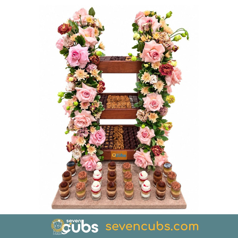 A decorative chocolate and pastry display stand shaped like the letter H, adorned with pink and cream roses and green foliage, holding multiple trays of chocolates and small dessert cups, on a white background with a teal brand banner at the bottom.